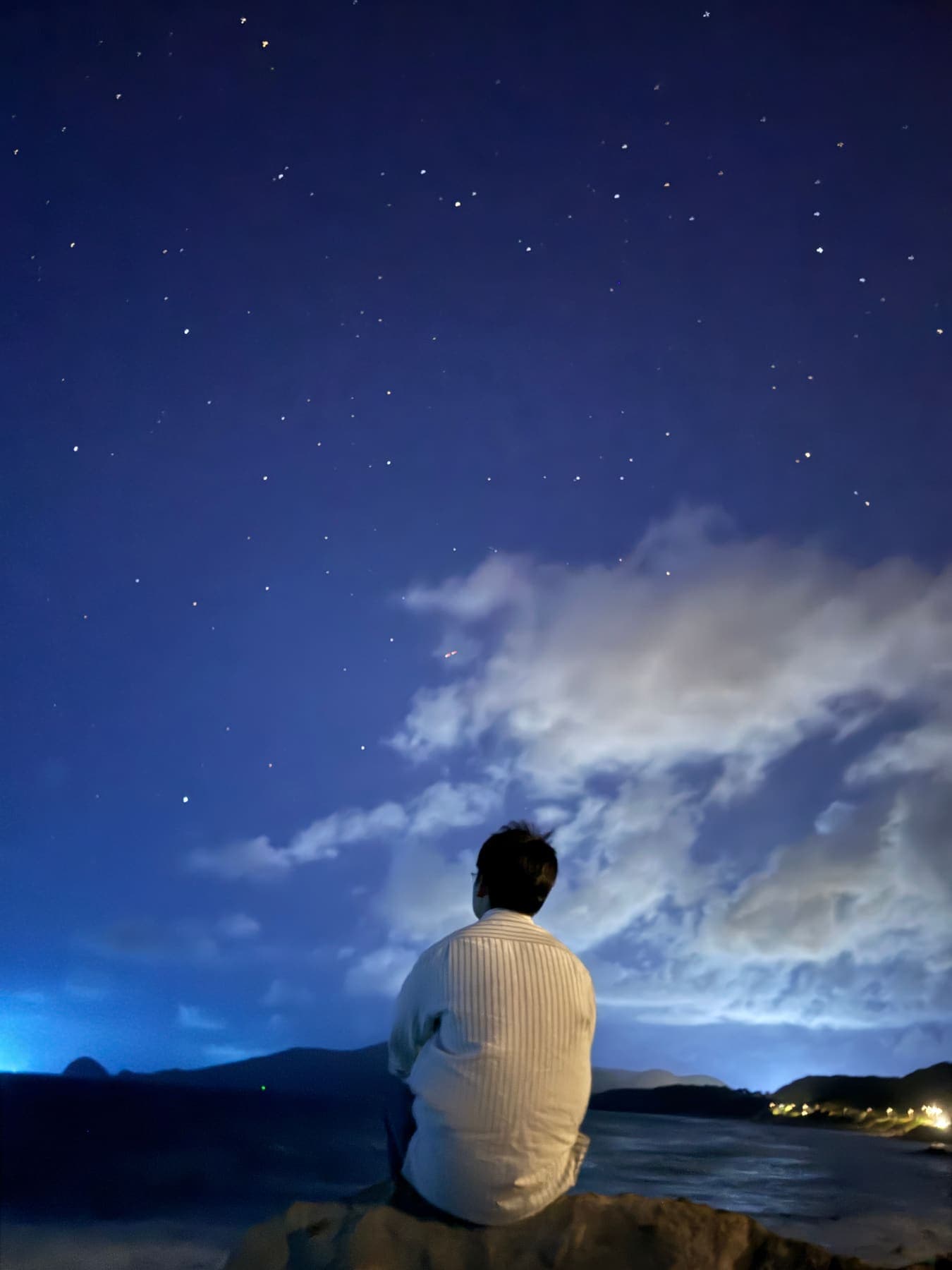 A man seen from behind on a rock, facing the sea under a starry sky