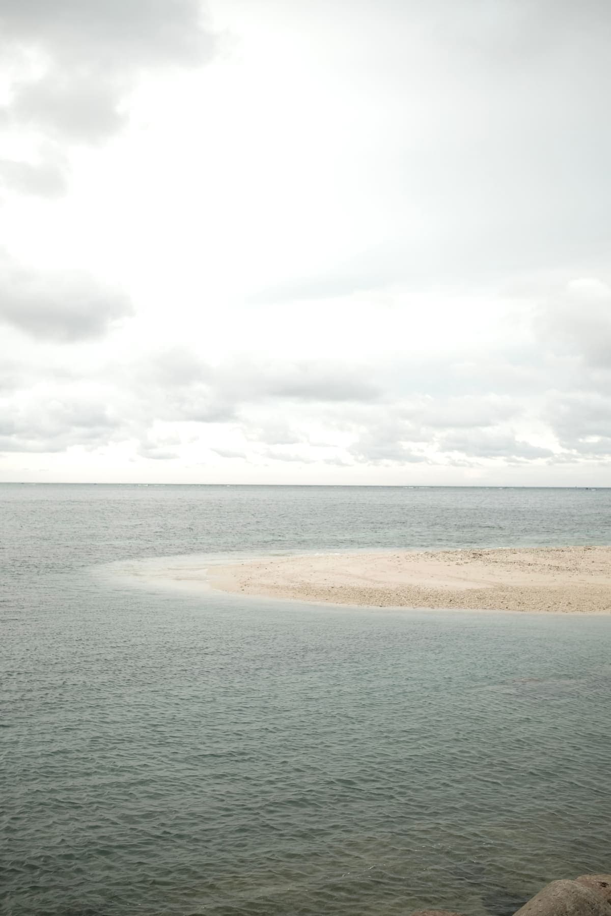 A pale sandbar curving into a calm grey sea under clouds