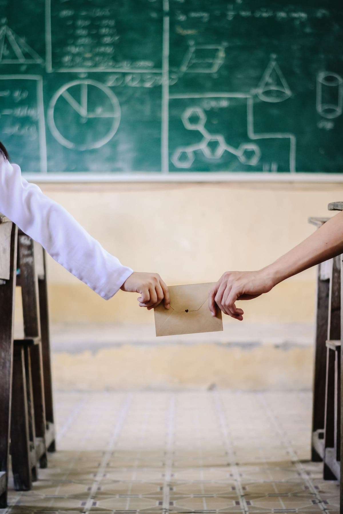 Two hands passing a folded note between desks in front of the blackboard