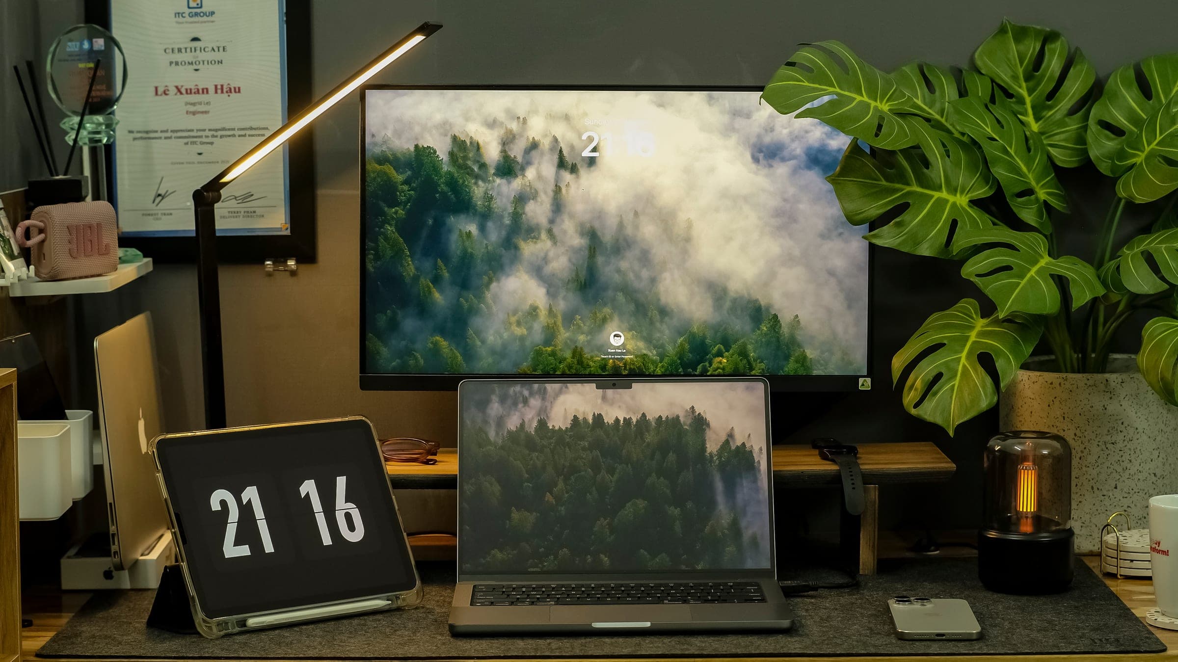 A desk setup at night: monitor with a misty forest wallpaper, laptop, flip clock and a monstera